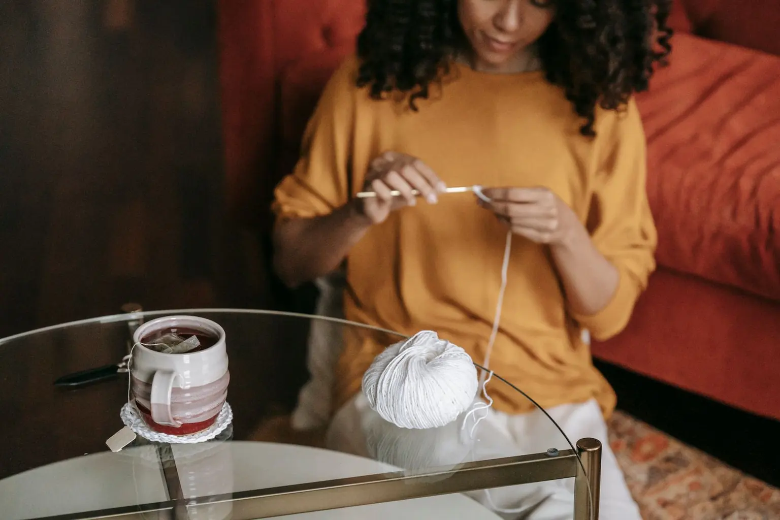 Mujer sentada en su sala teje con ganchillo e hilo blanco, con una taza de té y un ovillo sobre la mesa